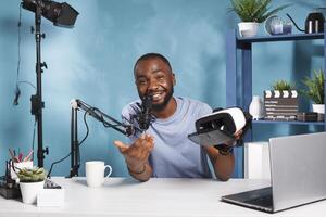 Cheerful african american blogger holding and showing vr headset in video presentation. Web content creator live streaming virtual reality glasses review and speaking in microphone photo