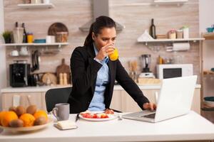 dama comiendo desayuno y utilizando ordenador portátil sentado en el silla en el cocina. concentrado negocio mujer en el Mañana multitarea en el cocina antes de yendo a el oficina, Estresante camino de vida, carrera y metas a encontrarse. foto