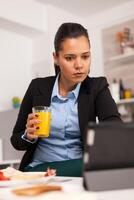 Young businesswoman holding a glass of juice and using the tablet in the morning. Freelancer reading the last news online before going to work, using modern technology in the kitchen while eating a healthy meal photo