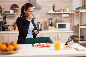 Busines woman eating breakfast and watching on tablet at home, in the kitchen. Business woman reading the last news online before going to work, using modern technology in the kitchen while eating a healthy meal. photo