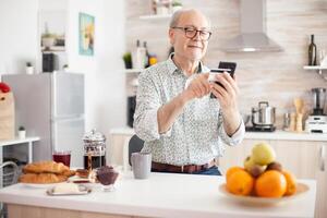 Senior man doing online transaction using phone app for payment during breakfast in kitchen. Retired elderly person using internet payment home bank buying with modern technology photo