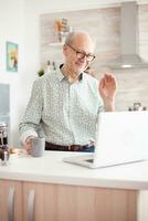 antiguo hombre diciendo Hola durante vídeo llamada con familia utilizando cuaderno en cocina mientras participación un taza de café. foto