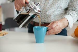 Senior man pouring hot coffee from french press during breakfat in kitchen. Elderly person in the morning enjoying fresh brown cafe espresso cup caffeine from vintage mug, filter relax refreshment photo