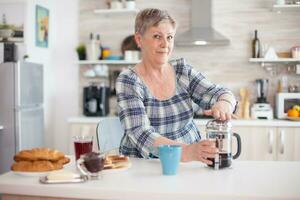 Portrait of old lady looking at camera while using french press in kitchen during breakfast. Elderly person in the morning enjoying fresh brown cafe espresso cup caffeine from vintage mug, filter relax refreshment photo
