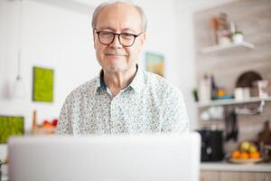 retirado hombre sonriente mientras acecho un película en el ordenador portátil. diario vida de mayor hombre en cocina durante desayuno utilizando ordenador portátil participación un taza de café. mayor retirado persona trabajando desde hogar, teletrabajo utilizando remoto Internet trabajo en línea comunicación. foto