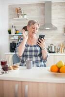 Retired woman saying hello while having a video call with family using smartphone in kitchen during breakfast. Elderly person using internet online chat technology video webcam making video call connection camera communication conference call photo