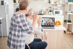 Happy senior woman waving during a video conference with family using laptop in kitchen. Online call with daughter and niece. Elderly person using modern communication online internet web techonolgy. photo