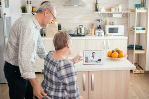 Senior couple listening doctor during telemedicine conference. Video conference with doctor using laptop in kitchen. Online health consultation for elderly people drugs ilness advice on symptoms, physician telemedicine webcam. Medical care internet chat photo