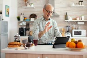 maduro hombre ondulación mientras teniendo un conversacion durante vídeo llamada en cocina disfrutando desayuno. mayor persona utilizando Internet en línea charla tecnología, tableta cámara web para virtual conferencia llamada foto