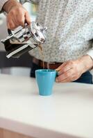 cerca arriba de mayor hombre derramar caliente bebida después preparando café utilizando francés prensa en cocina para desayuno. mayor persona en el Mañana disfrutando Fresco marrón café Café exprés taza cafeína desde jarra foto