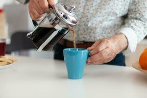 Close up of senior man pouring hot coffee from french press in kitchen during breakfast. Elderly person in the morning enjoying fresh brown cafe espresso cup caffeine from vintage mug, filter relax refreshment photo