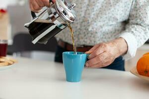 retirado hombre participación azul taza en cocina mesa torrencial caliente aromático bebida desde un café máquina durante desayuno. mayor persona en el Mañana Bebiendo Fresco marrón Café exprés desde Clásico jarra foto