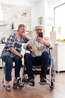 Smiling senior woman and her disabled husband in wheelchair using smartphone in kitchen, surfing on social media in the morning. paralyzedhandicapped old man using modern communication techonolgy. photo