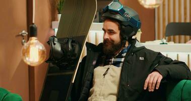 Athletic traveler seated in lobby looking at his snowboard and feeling excited to start winter sport on mountain slopes. Young man equipped with helmet and goggles ready for wintertime leisure. photo