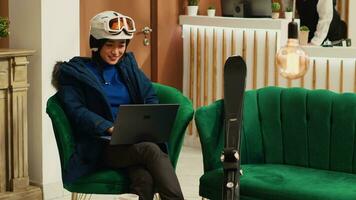 Tourist reading reviews on laptop at luxury hotel, sitting in lounge area with skiing equipment and accessories. Asian woman working remote on pc before practicing extreme winter sport in the cold. photo