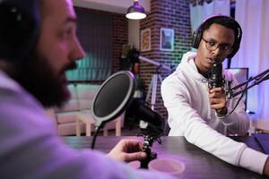 Wise guest on online podcast show telling inspiring story to host, entertaining viewers watching during live broadcast. Presenter using audio recording equipment in rgb lights decorated living room photo