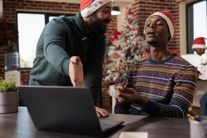 Company team lead in santa hat helping employee with project while working in christmas decorated office. Manager pointing on laptop and explaining task to coworker in festive workplace photo
