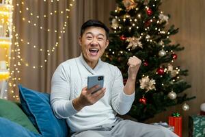 A young Asian man sits at home on the sofa near the Christmas tree, holds the phone in his hands, rejoices at the Christmas win in the raffle, the lottery prize, received good news, a message. photo
