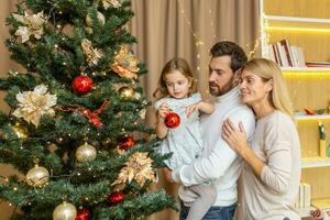 A young happy family decorates a Christmas tree for the Christmas holidays. Dad holds his daughter in his arms, the girl hangs a toy, mom hugs dad. photo