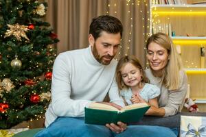 A young happy family spends time together on Christmas Eve. Mom and dad are reading a book to their daughter, sitting on the sofa near the Christmas tree. photo