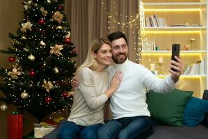 casado Pareja hombre y mujer juntos en sofá a hogar Navidad árbol con teléfono inteligente hablando vídeo llamada y tomando autofoto, maduro familia saludo amigos en línea remotamente para nuevo año y Navidad. foto