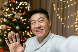 contento asiático hombre celebrando Navidad y nuevo año solo a hogar, hombre mirando a teléfono inteligente cámara y sonriente, hablando con amigos en ,teléfono vídeo llamada atractivo invitados mientras, sentado en sofá foto