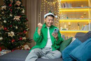 A young Asian man is sitting at home near the Christmas tree, in a red hat, waiting for guests for the New Year's party. Plays games on the phone. Happy won the game, shows yes with his hand. photo