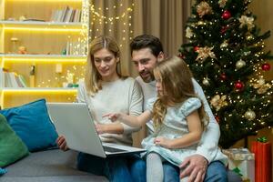 un niño niña elige un regalo para Navidad en un ordenador portátil en línea, puntos a el monitor. sentado con mamá y papá en el sofá a hogar cerca el Navidad árbol. foto