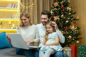 Happy family home for Christmas, husband wife and little daughter sitting on sofa in living room near Christmas tree, looking laptop and smiling celebrating New Year holidays, choosing gifts online. photo