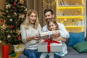 festivo retrato de un joven contento familia cerca el Navidad árbol a hogar. mamá, papá y bebé hija son sentado en el sofá y intercambiando regalos, alegremente dando cada otro cajas con sorpresas foto