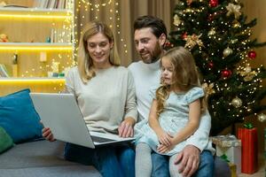 Happy family man wife and daughter celebrating new year and Christmas together, couple sitting on sofa at home near Christmas tree with laptop, watching video online together. photo
