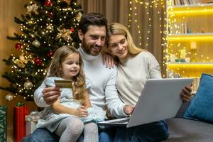 Christmas family, husband wife and little daughter for Christmas at home make online shopping in online store remotely, smiling and happy, husband holding bank credit card and laptop choosing gifts. photo