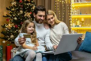 Navidad familia, marido esposa y pequeño hija para Navidad a hogar hacer en línea compras en en línea Tienda de forma remota, sonriente y feliz, marido participación banco crédito tarjeta y ordenador portátil elegir regalos. foto