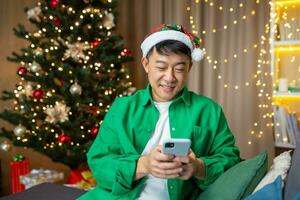 A young Asian man in a red Santa Claus hat and a green shirt holds a phone in his hands, writes a message, congratulates on the Christmas holidays. Sitting at home on the sofa near the Christmas tree. photo