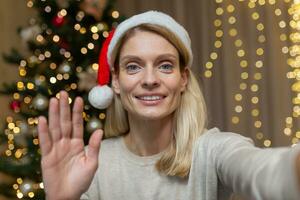 maduro mujer a hogar celebrando nuevo año y Navidad, rubia hablando en vídeo llamada utilizando aplicación en teléfono a comunicar con amigos de forma remota, esposa cerca Navidad árbol en vivo habitación a hogar. foto