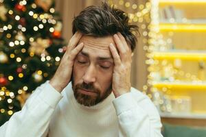 un hombre tiene un dolor de cabeza para Navidad, un de cerca foto en nuevo años víspera cerca el árbol, un hombre participación su cabeza con su manos, enfermo, sentado en el sofá a hogar solo.