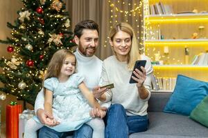 Young family doing holiday shopping online. A woman holds a credit card and a phone in her hands. She shows her husband and daughter. They are sitting at home on the sofa near the Christmas tree. photo
