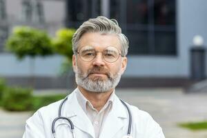 Closeup portrait of mature man in medical coat, senior doctor thinking and looking at camera near clinic outside. photo