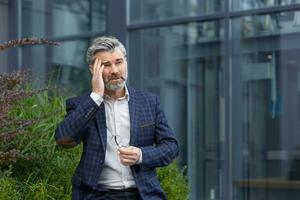 Mature fairhaired man outside office building has dizziness and headache, businessman in business suit is overwhelmed in the air, senior boss is sick. photo