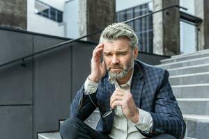 Gray haired businessman sitting sad on stairs outside office building, depressed boss in business suit upset and disappointed with achievement results. photo