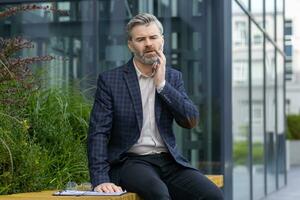 Toothache, mature adult male businessman sitting outside office building, having severe pain, holding hand to face, gingivitis in gray haired boss. photo