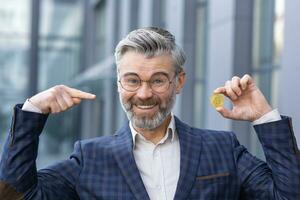 retrato de un contento mayor empresario en pie en un traje fuera de un oficina centrar y participación bitcoin, criptomoneda en uno mano, señalando a el moneda con el otro mano. foto