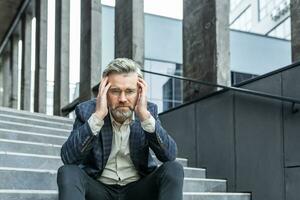 Gray haired businessman sitting sad on stairs outside office building, depressed boss in business suit upset and disappointed with achievement results. photo