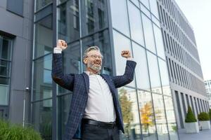 Successful and happy grayhaired boss outside office building rejoices success and victory, triumphs with hands up, businessman business owner in business suit and glasses, mature investor banker photo