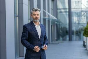 Portrait of successful mature man outside office building, businessman in business suit looking at camera thinking smiling while standing. photo