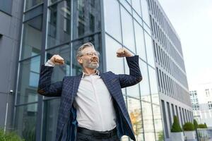 Successful and happy grayhaired boss outside office building rejoices success and victory, triumphs with hands up, businessman business owner in business suit and glasses, mature investor banker. photo