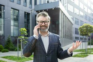 Portrait of happy mature businessman, gray haired man outside office building talking on the phone, boss smiling and talking to colleagues. photo