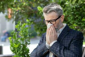 Sick and cold mature adult man sitting on bench outside office building, businessman in business suit sneezing and having runny nose seasonal allergy, employee boss during work break on bench. photo