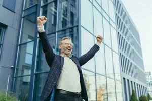 Successful and happy grayhaired boss outside office building rejoices success and victory, triumphs with hands up, businessman business owner in business suit and glasses, mature investor banker. photo