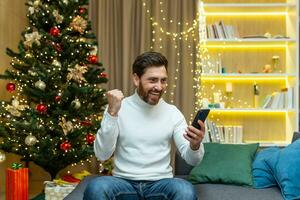 Man celebrating victory and success received good notification online message sitting on sofa near tree during christmas and new year, holidays holding hand up success gesture using phone. photo